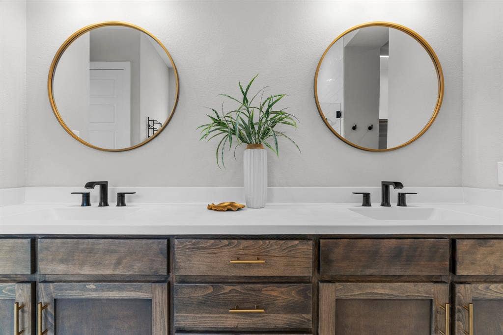 1821 Stonewall Road, Argyle, TX 76226 Dual vanity featuring a light-toned countertop, two round gold-framed mirrors, and dark wood-finish cabinetry with gold hardware