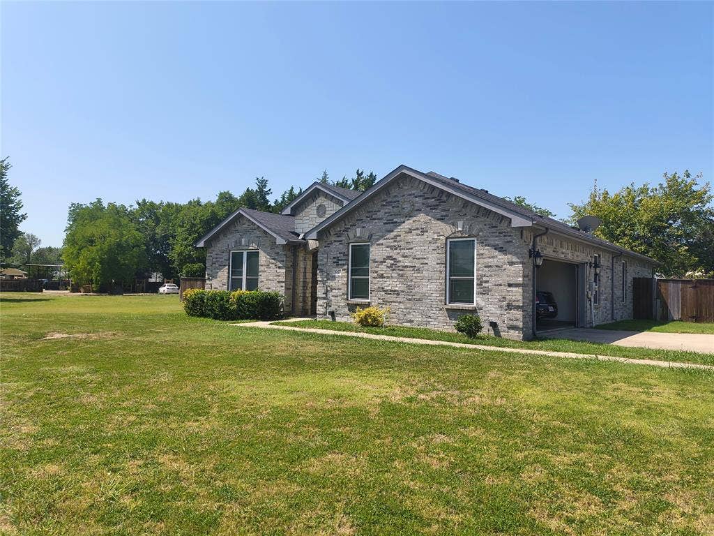 3027 Ames Road, Lancaster, TX 75134 Grey brick exterior with multiple window openings, an attached garage, a dark asphalt shingle roof, a concrete driveway, and front landscaping