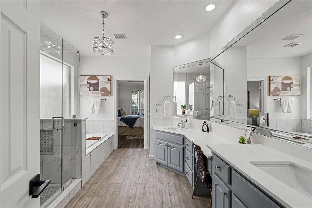 7904 Alderwood Place, Plano, TX 75025 Spacious bathroom featuring a frameless glass shower enclosure, a built-in soaking tub, wood-finish flooring, dual vanities with light gray cabinetry, and an expansive wall-mounted mirror