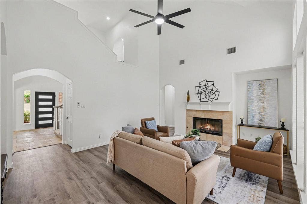 7904 Alderwood Place, Plano, TX 75025 Spacious living room featuring vaulted ceilings, a modern ceiling fan, wood-finish flooring, a tiled fireplace with mantel, and an arched entry to the foyer