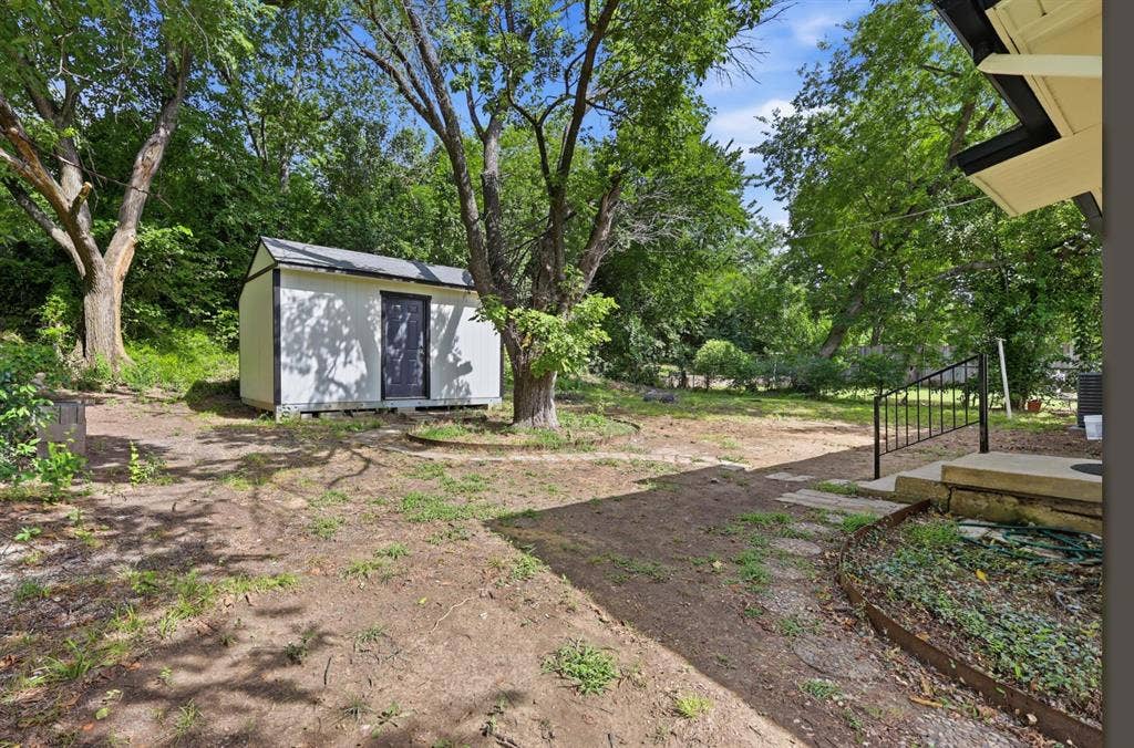 810 N Carrier Parkway, Grand Prairie, TX 75050 Spacious backyard featuring a white storage shed with dark trim, mature trees, a concrete patio with black metal railing, and stepping stones