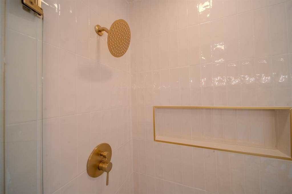 Shower enclosure featuring white subway tile, a gold-finish rainfall showerhead, coordinating gold-finish controls, a glass partition, and an integrated recessed niche with gold trim