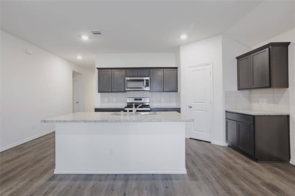 2904 Goodrich Street, Royse City, TX 75189 Open-concept kitchen featuring an island with a light-toned countertop, dark wood-finish cabinetry, stainless steel appliances, a white tile backsplash, and wood-finish flooring