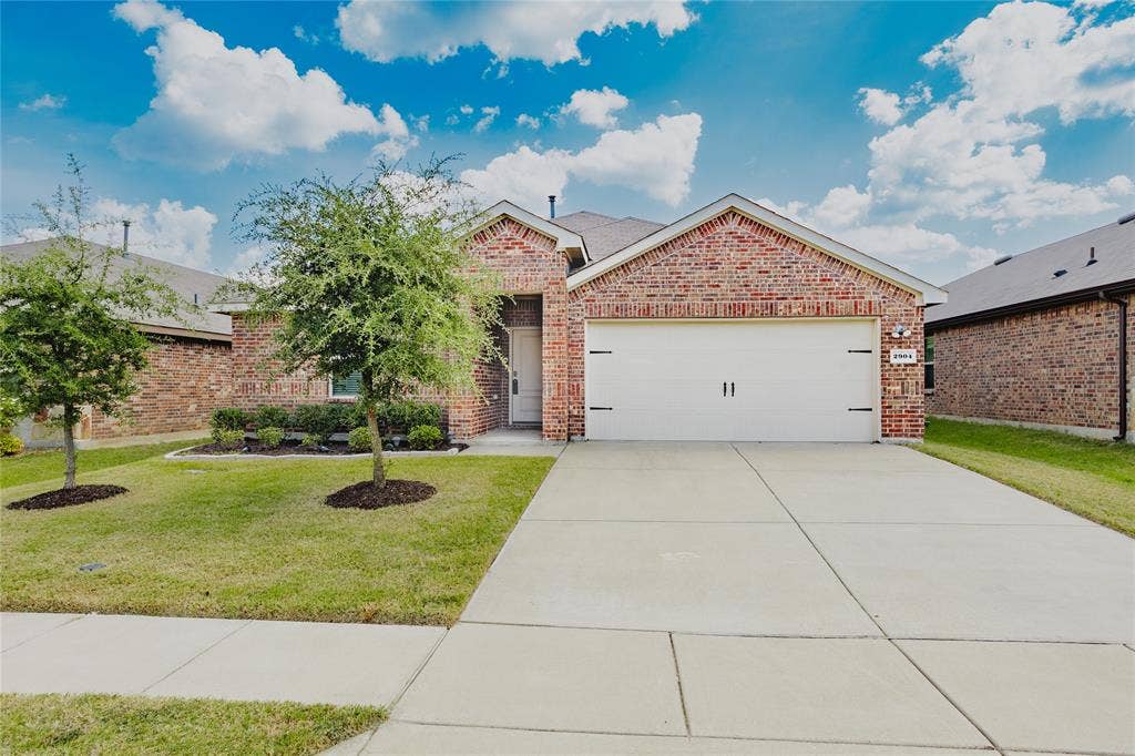 2904 Goodrich Street, Royse City, TX 75189 Brick exterior featuring a two-car garage with decorative hardware, an extended concrete driveway, and a manicured lawn with mature trees