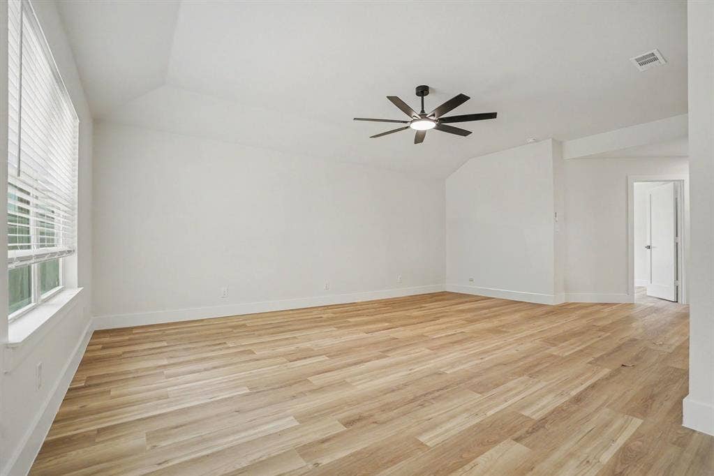 Spacious bedroom with warm wood-finish flooring throughout, abundant natural light from large windows with plantation shutters, and a modern ceiling fan with integrated lighting fixture