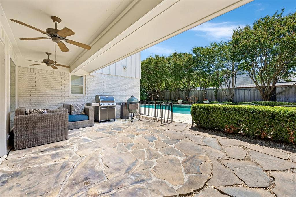 4110 Shady Hill Drive, Dallas, TX 75229 A covered flagstone patio with ceiling fans overlooks the pool and landscaped backyard, creating an inviting space for outdoor living and entertaining.