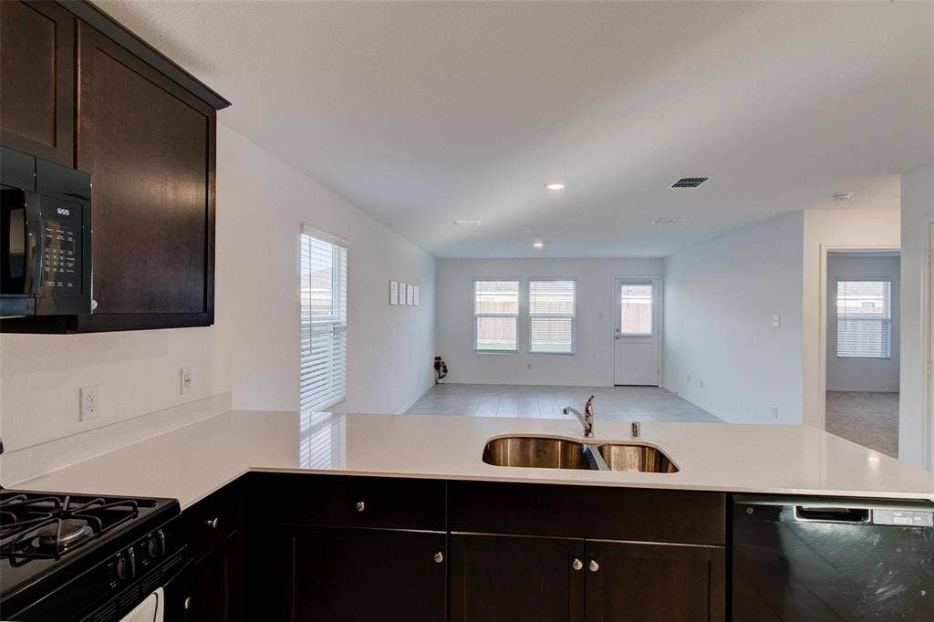 6005 Determine Lane, Forney, TX 75126 Kitchen featuring dark wood cabinetry, light-toned countertops, a built-in stainless steel double sink, and integrated black appliances including a microwave, gas range, and dishwasher