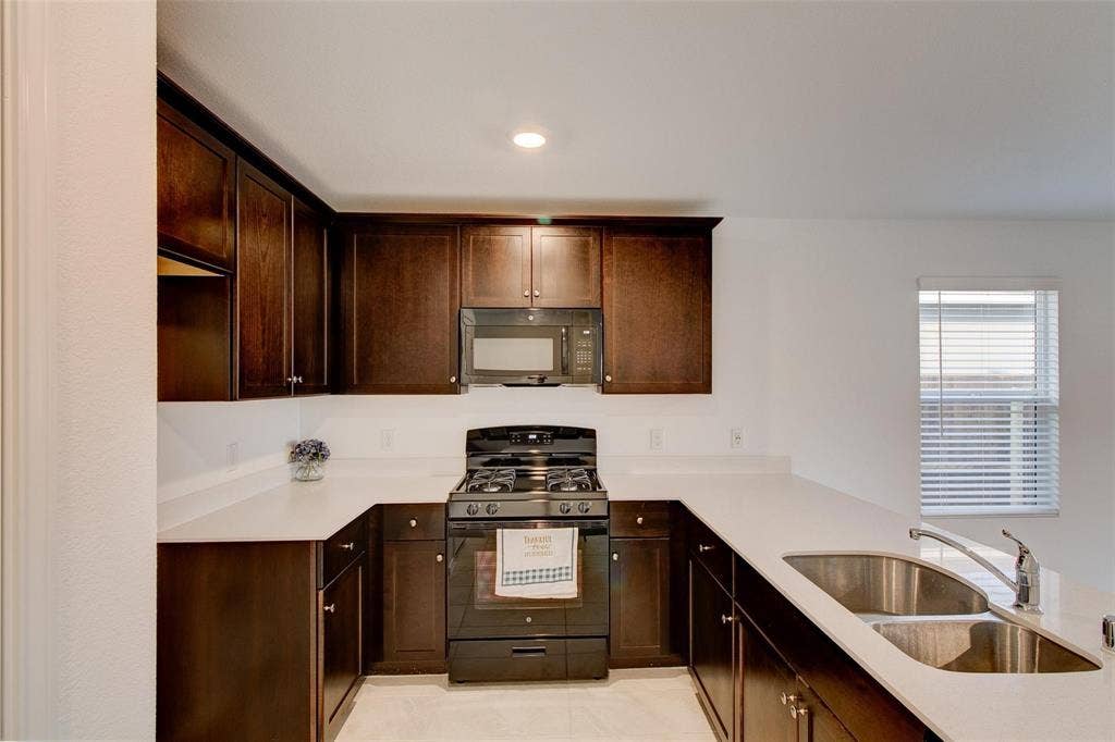 6005 Determine Lane, Forney, TX 75126 U-shaped kitchen featuring dark wood cabinetry, light countertops, a double basin stainless steel sink with a gooseneck faucet, a built-in microwave, and a gas range