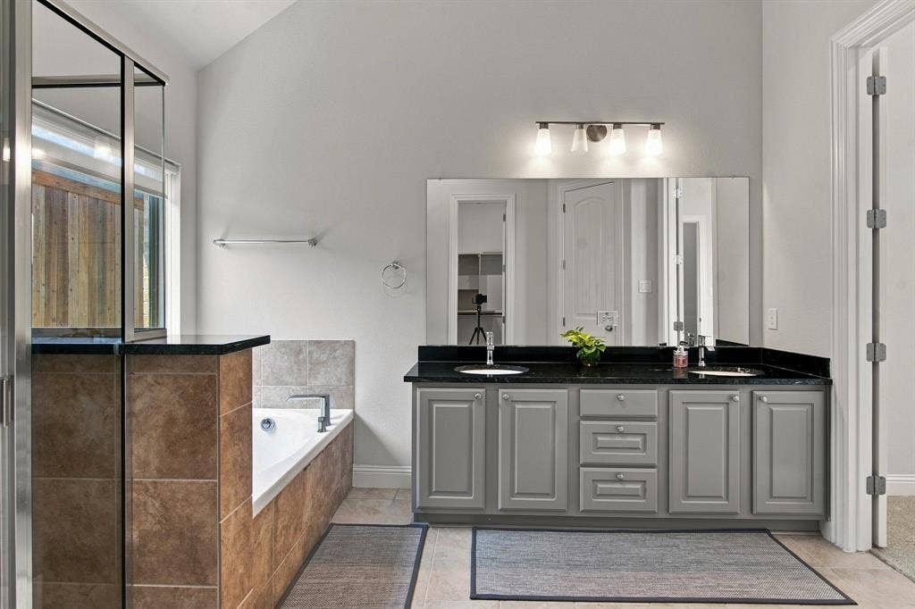 944 Tara Court, Haslet, TX 76052 Spa-inspired primary bathroom featuring a dual-sink vanity with gray cabinetry, black countertops, and polished hardware, complemented by a frameless glass-enclosed shower with black accents, large mirror with integrated lighting, and neutral tile flooring