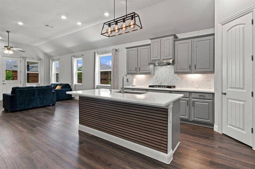 944 Tara Court, Haslet, TX 76052 Open-concept kitchen and living area featuring wood-finish flooring, vaulted ceilings, a central kitchen island with a slatted wood-look accent, gray cabinetry, and white countertops with a tiled backsplash
