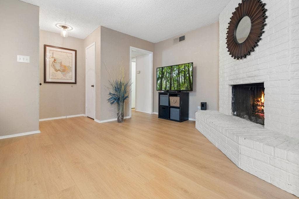 Expansive living area featuring light wood-finish flooring, a white-painted brick fireplace with a hearth, and neutral wall tones