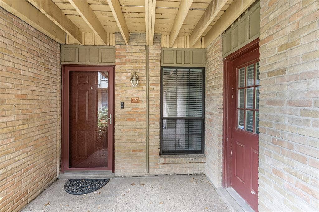 5916 Birchbrook Drive, #129, Dallas, TX 75206 Covered entryway featuring brick facade, red-paneled entry door with glass inserts, transom window, and exposed wood beam ceiling
