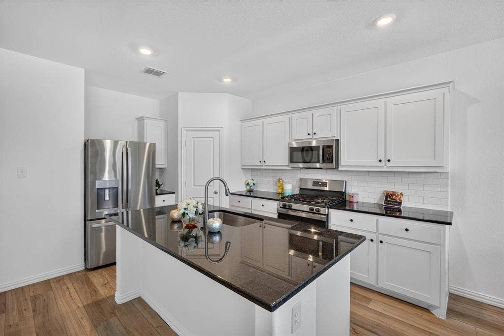 Modern kitchen featuring white shaker-style cabinetry, black quartz countertops, a large waterfall-edge island with seating, stainless steel appliances including a built-in microwave and gas cooktop, recessed lighting, and wood-finish flooring throughout