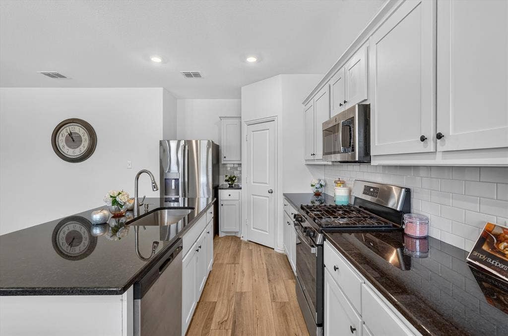 Modern kitchen featuring white shaker cabinetry, dark countertops, a stainless steel gas range, microwave, and refrigerator, complemented by wood-finish flooring and subway tile backsplash