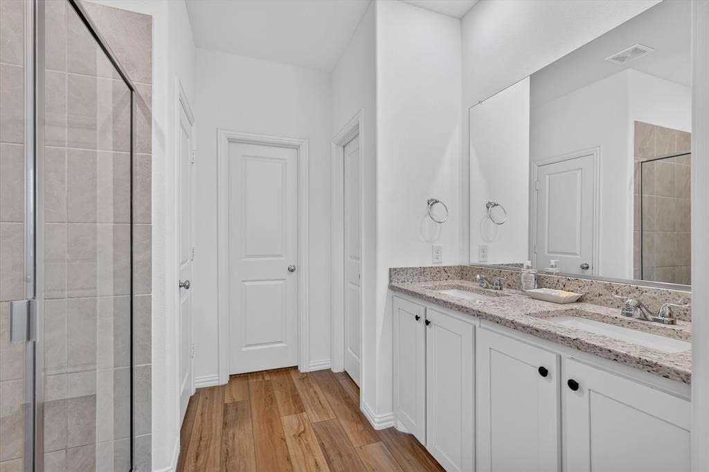 Double vanity with undermount sinks and speckled countertops, frameless mirror, wood-finish flooring, and a glass-enclosed shower with light-toned tile