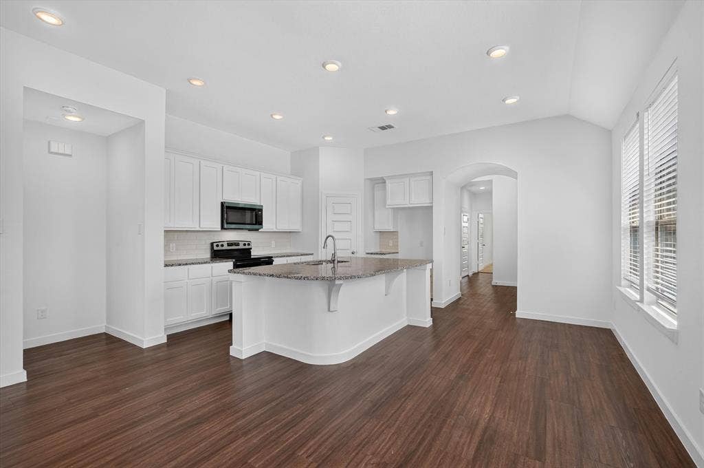2832 Lolita Way, Forney, TX 75126 Modern kitchen and dining area featuring white cabinetry with stainless steel appliances, a curved island with dark countertop and undermount sink, recessed ceiling lighting, wood-finish flooring throughout, and an arched passageway leading to additional living spaces