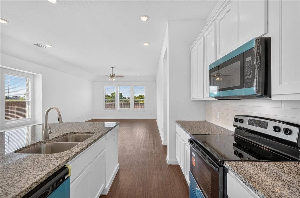 2832 Lolita Way, Forney, TX 75126 Modern galley kitchen featuring white shaker cabinetry, granite countertops, stainless steel appliances including a convection microwave and electric range, a dual-basin undermount sink with gooseneck faucet, wood-finish flooring, and abundant natural light from multi-pane windows overlooking the landscape