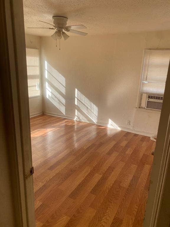 Wood-finish flooring, ceiling fan with integrated lighting, window-mounted air conditioning unit, and multiple windows with blinds