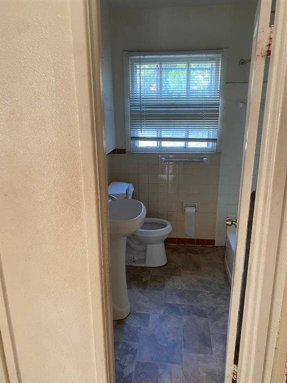 Pedestal sink, window with blinds, wall-mounted toilet paper holder, ceramic wall tile, and a bathtub