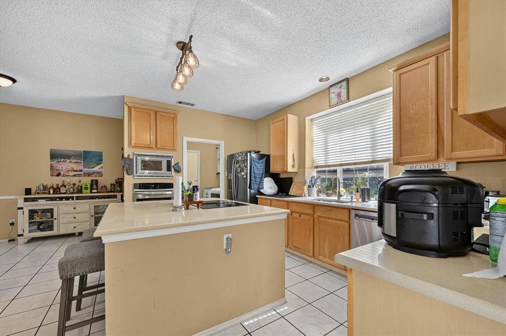 1625 Falmouth Drive, Plano, TX 75025 Kitchen featuring light wood cabinetry, white tile flooring, a central island with a light-toned countertop, stainless steel appliances, and a window with horizontal blinds