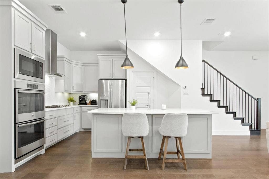 7867 Verona Place, Dallas, TX 75231 Modern kitchen featuring light cabinetry, stainless steel appliances, a center island with pendant lighting, wood-finish flooring, and a contemporary staircase with dark metal balusters