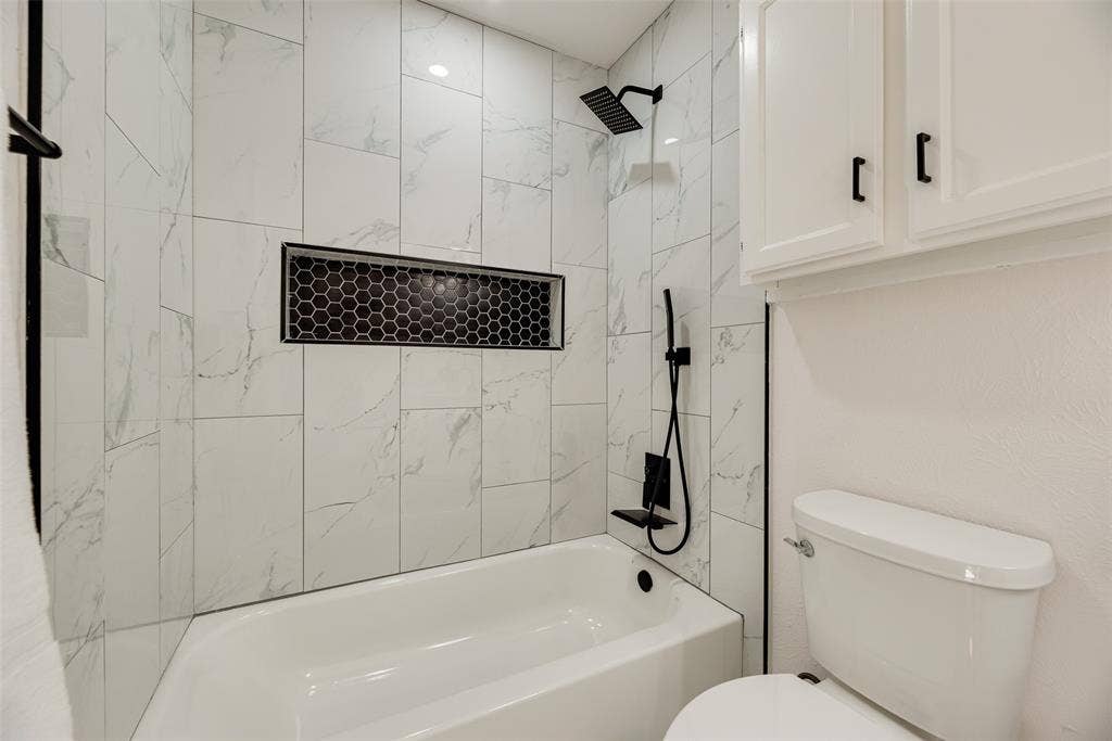 Bathtub and shower combination featuring large format white tile with grey veining, a black hexagonal tile shower niche, and matte black shower fixtures