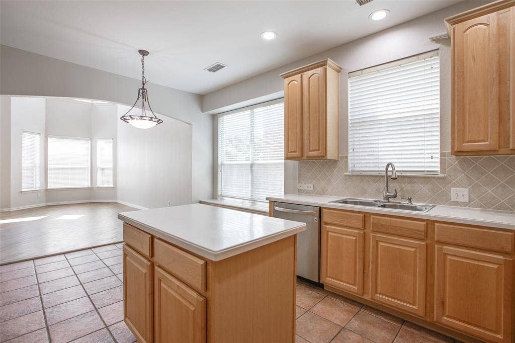 3401 Maddock Drive, McKinney, TX 75070 Spacious kitchen featuring warm wood cabinetry with raised-panel doors, a large center island with white quartz countertop, stainless steel sink with faucet, tile flooring, and abundant natural light from multiple windows with blinds