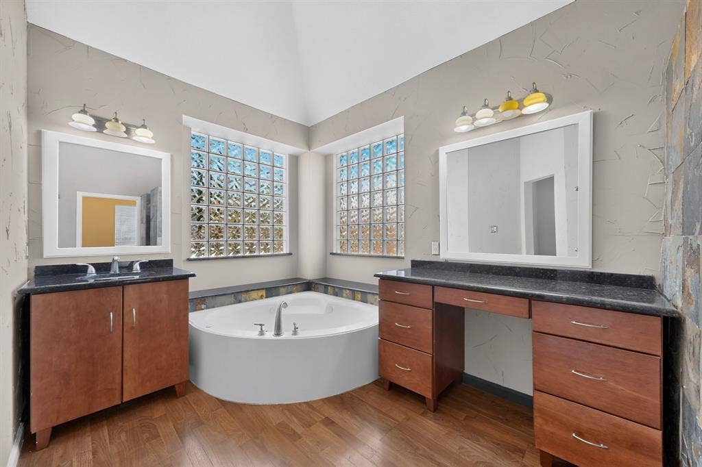 713 Mockingbird Court, Crowley, TX 76036 Soaking tub with deck-mounted fixtures, dual vanity sinks with dark countertops, wood-finish flooring, and glass block windows
