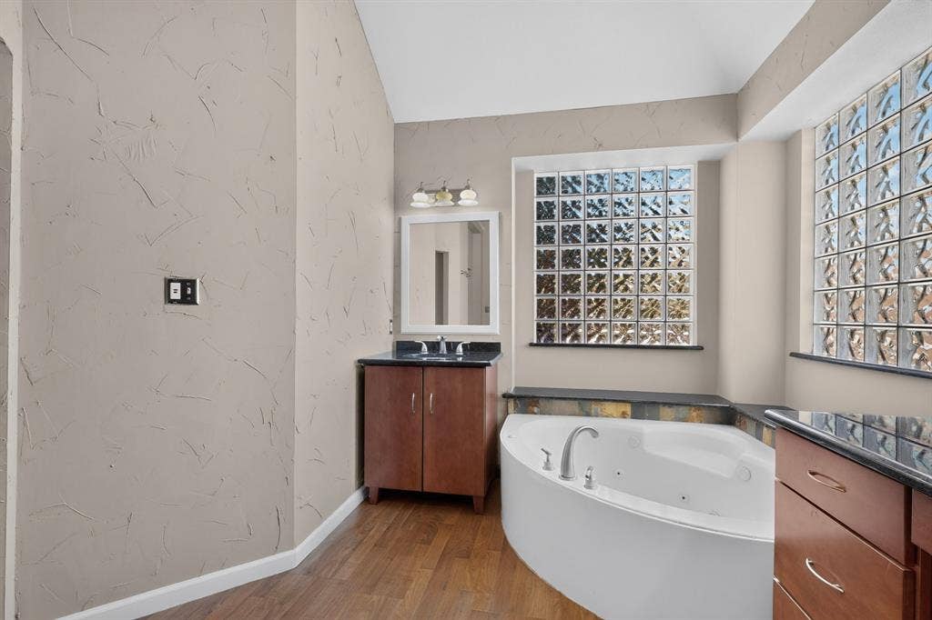 713 Mockingbird Court, Crowley, TX 76036 Soaking tub with deck-mounted fixtures, two glass block windows, slate-style tile surround, wood-finish flooring, and a vanity with dark countertop