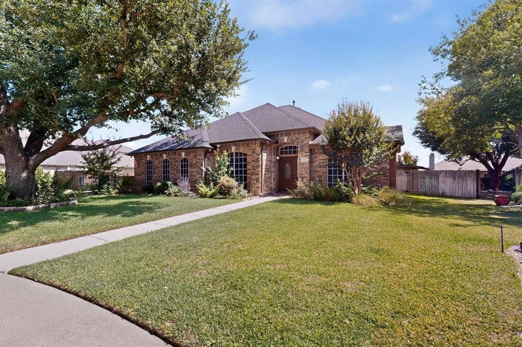 713 Mockingbird Court, Crowley, TX 76036 Brick exterior featuring arched windows, a dark shingle roof, mature trees, and a concrete walkway