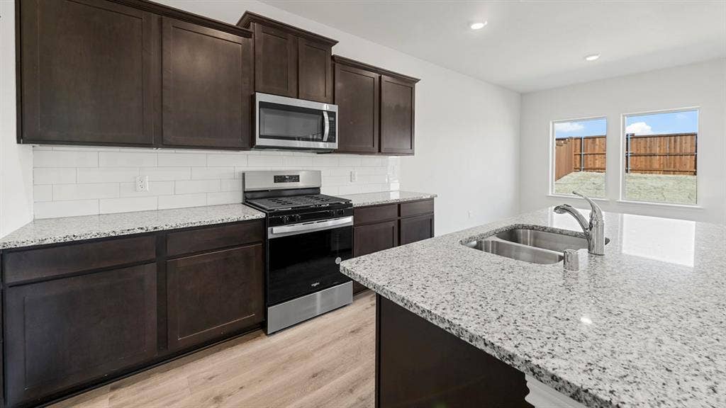 8425 Everly Drive, Garland, TX 75043 Modern kitchen featuring dark cabinetry, stainless steel gas range and microwave, granite-style countertops with waterfall edge, stainless steel single-basin sink with chrome faucet, subway tile backsplash, and wood-finish flooring throughout