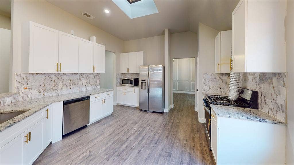3312 Woodberry Lane, McKinney, TX 75071 Galley-style kitchen featuring white cabinetry with brushed gold hardware, marble subway tile backsplash, stainless steel appliances including a gas range and refrigerator, quartz countertops, wood-finish flooring, and skylight overhead for natural illumination