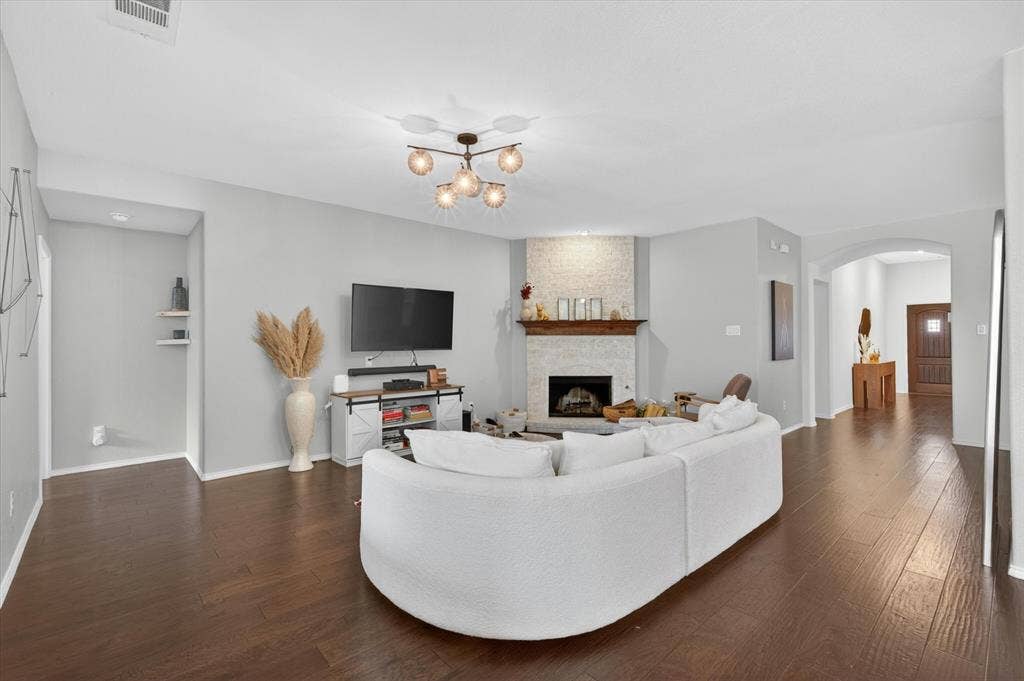 Spacious open-concept living area showcasing a curved white sectional sofa, dark wood-finish flooring throughout, and a gas fireplace with stone surround and mantel