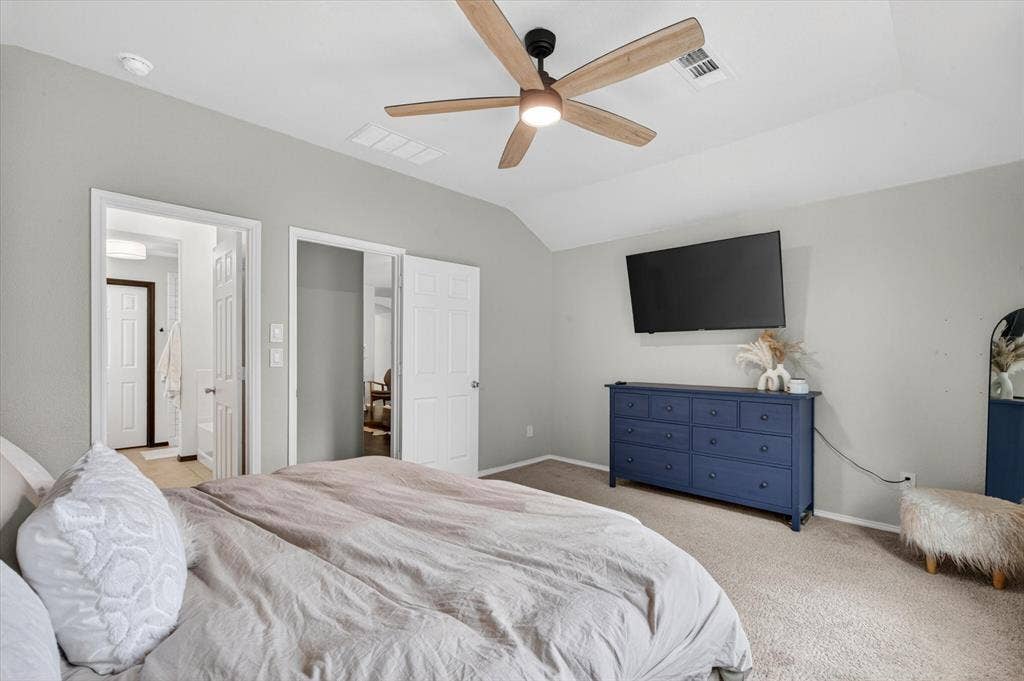 Spacious primary bedroom featuring neutral wall finishes, wood-finish ceiling fan with integrated lighting, wall-mounted flat-screen television, and direct access to an ensuite bathroom