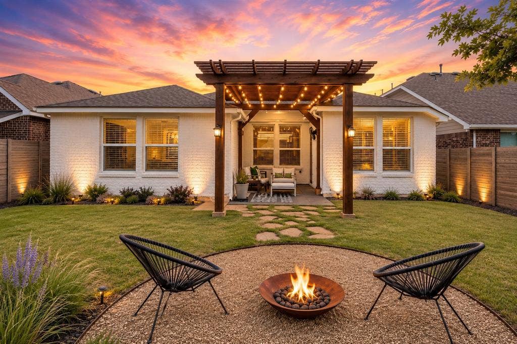 Manicured lawn with a stone-paved patio, a wood pergola with string lighting, and a gravel fire pit area