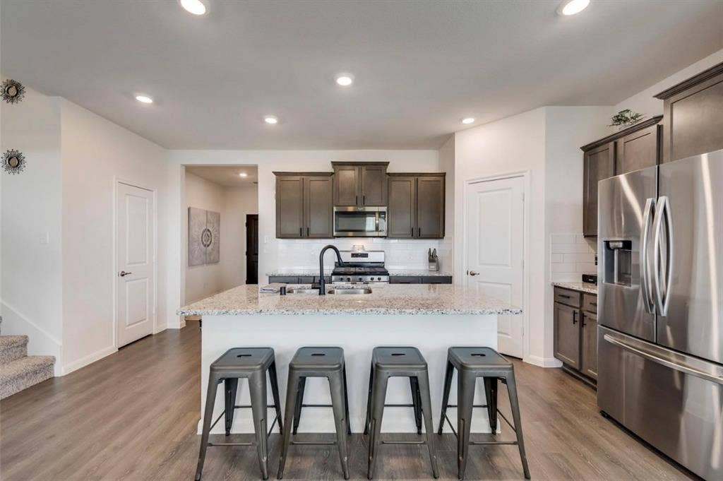 902 Western Way, Princeton, TX 75407 Modern kitchen featuring a spacious white quartz island with seating for four, stainless steel appliances including a gas cooktop and side-by-side refrigerator, dark cabinetry with integrated hardware, recessed ceiling lighting, wood-finish flooring, and an open layout connecting to the adjoining living spaces
