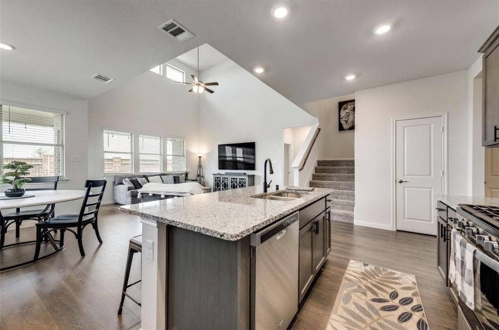 902 Western Way, Princeton, TX 75407 Open-concept kitchen and living area featuring a large center island with granite countertop and dark cabinetry, stainless steel appliances including a dual-fuel range, recessed and pendant lighting, vaulted ceilings with skylights, hardwood-finish flooring, and direct sightlines to a multi-level staircase and secondary living spaces