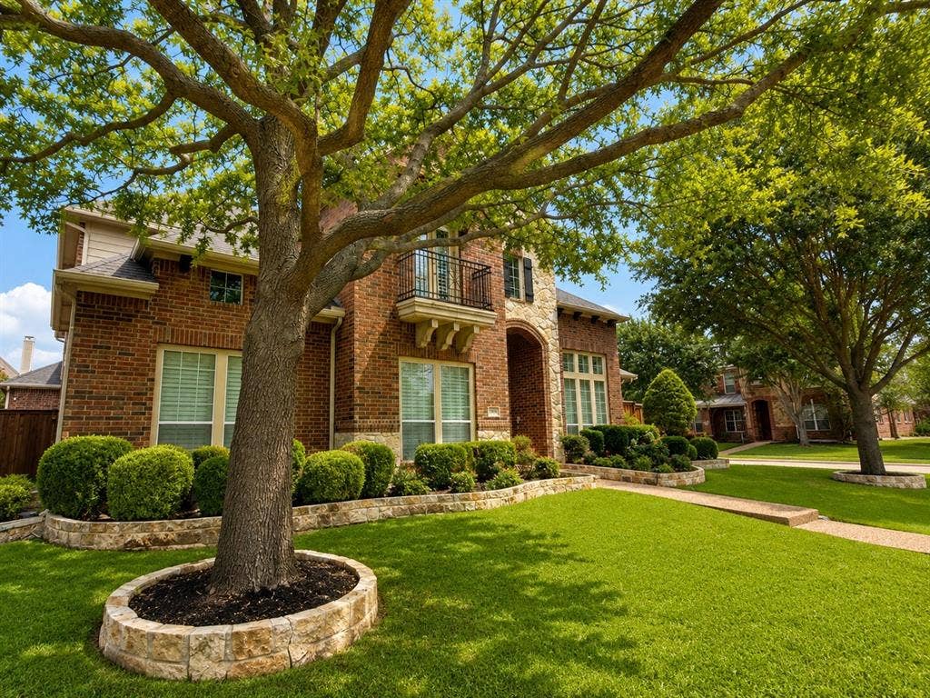 Brick and stone exterior featuring a second-story balcony with iron railing, manicured front lawn, and mature trees