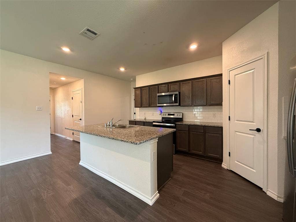 2702 Marotto Way, Seagoville, TX 75159 Kitchen featuring an island with a built-in sink, granite countertops, wood-finish flooring, dark wood cabinetry, and stainless steel appliances