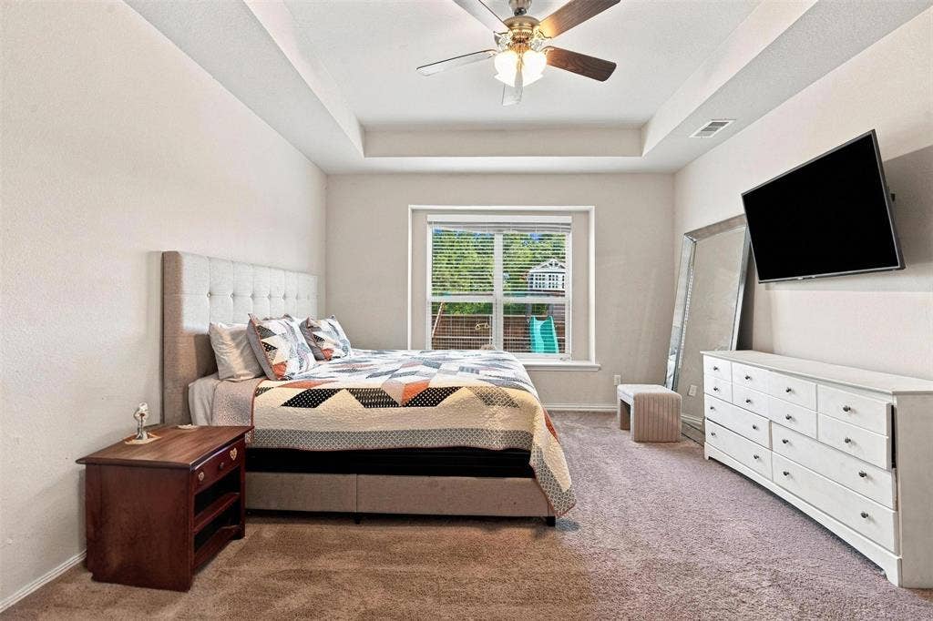 Bedroom featuring a tray ceiling with a ceiling fan, neutral carpet flooring, and a large window with horizontal blinds