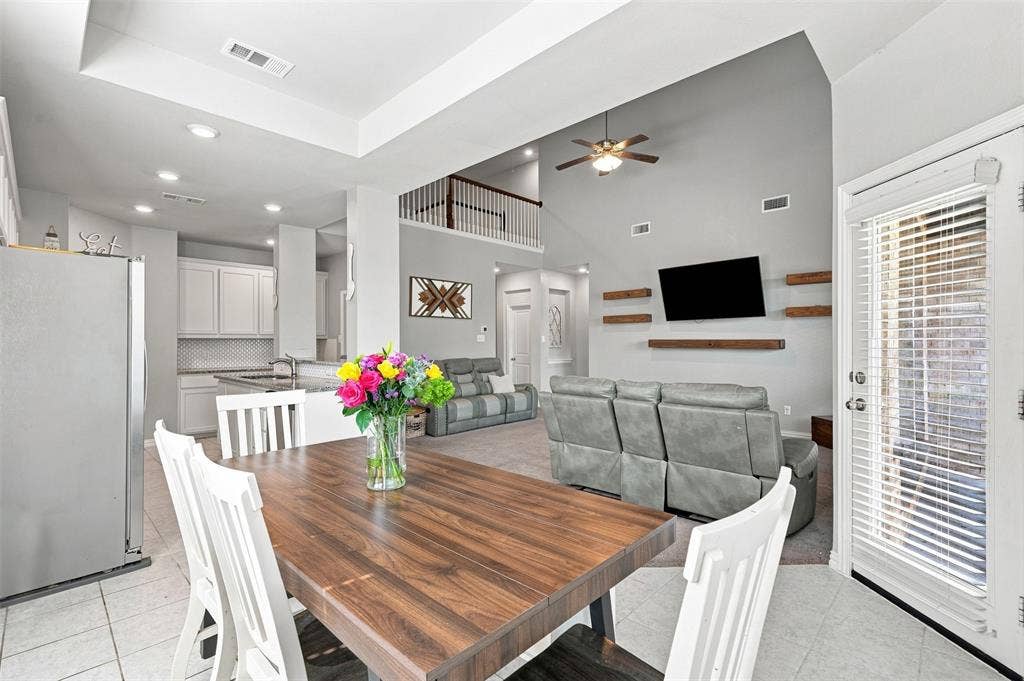 Open concept living space featuring a wood-finish dining table, light tile flooring, and a kitchen with white cabinetry and a mosaic tile backsplash