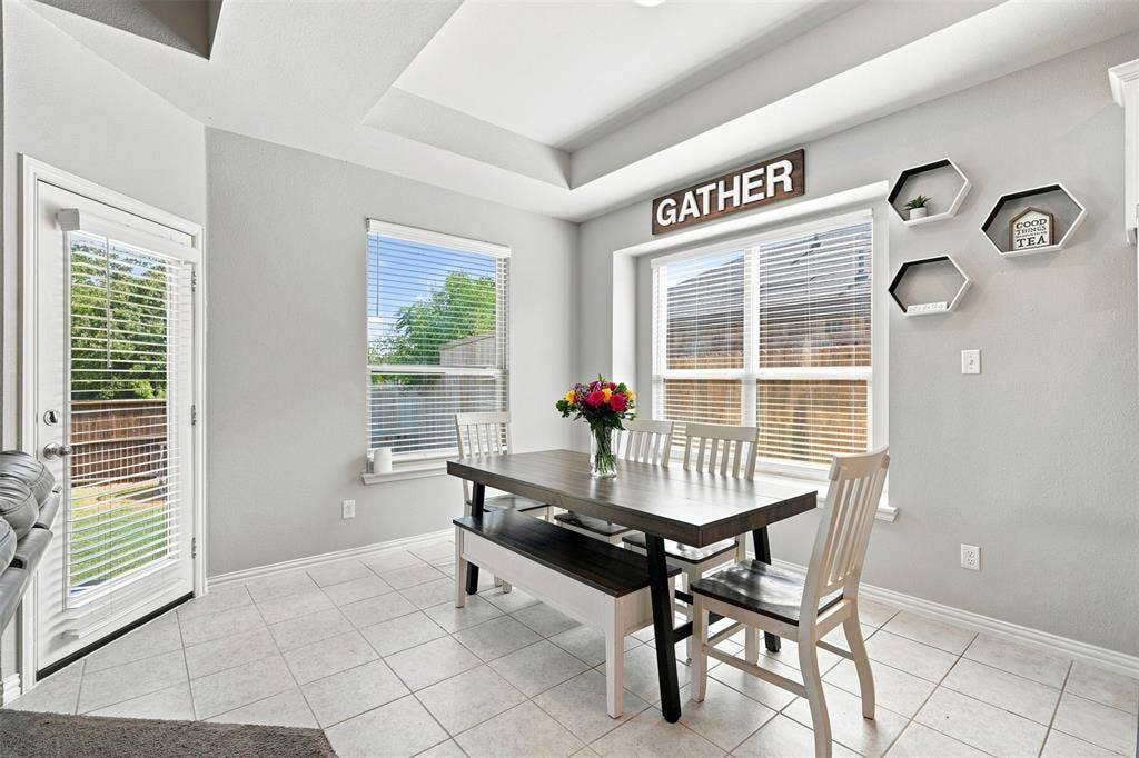 Bright dining area featuring a tray ceiling, tile flooring, two large windows with blinds, and a glass-paneled exterior door