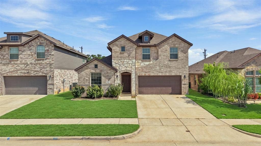 Two-story stone and brick facade residence featuring a two-car garage, dormer windows, and a landscaped front yard with a concrete driveway