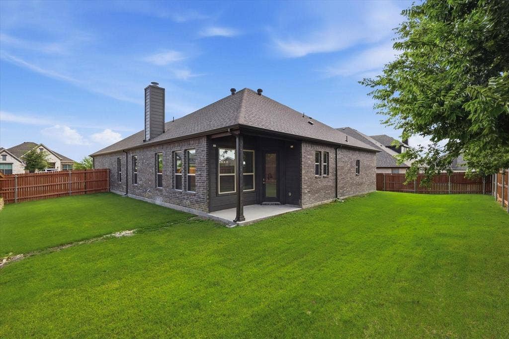 Generous green lawn encompassing the rear of the property, featuring a covered patio, dark brick exterior, multiple windows, and a privacy fence