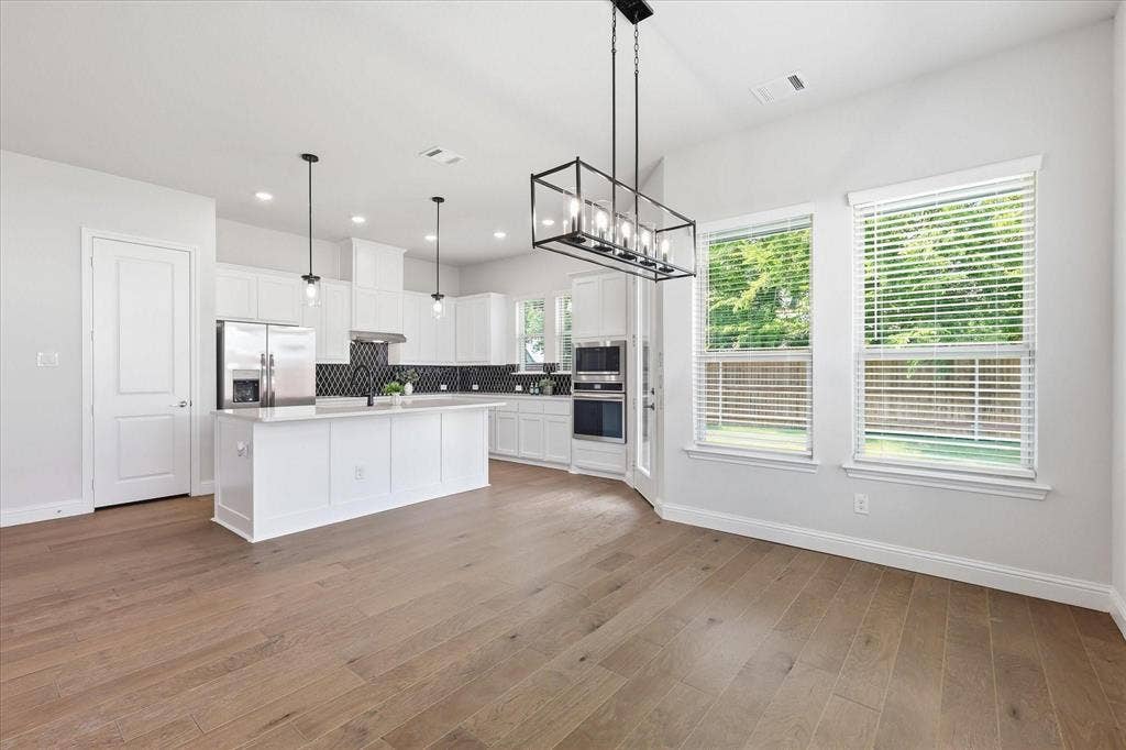 Open-concept kitchen and dining area featuring light wood-finish flooring, white cabinetry, stainless steel appliances, a kitchen island with pendant lighting, and a black geometric tile backsplash
