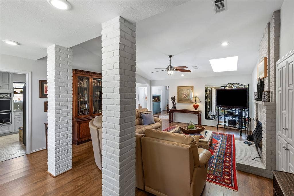 6803 Fernshaw Drive, Dallas, TX 75248 Vaulted ceiling living area featuring a skylight, wood-finish flooring, white-painted brick columns, and a brick fireplace with built-in cabinetry