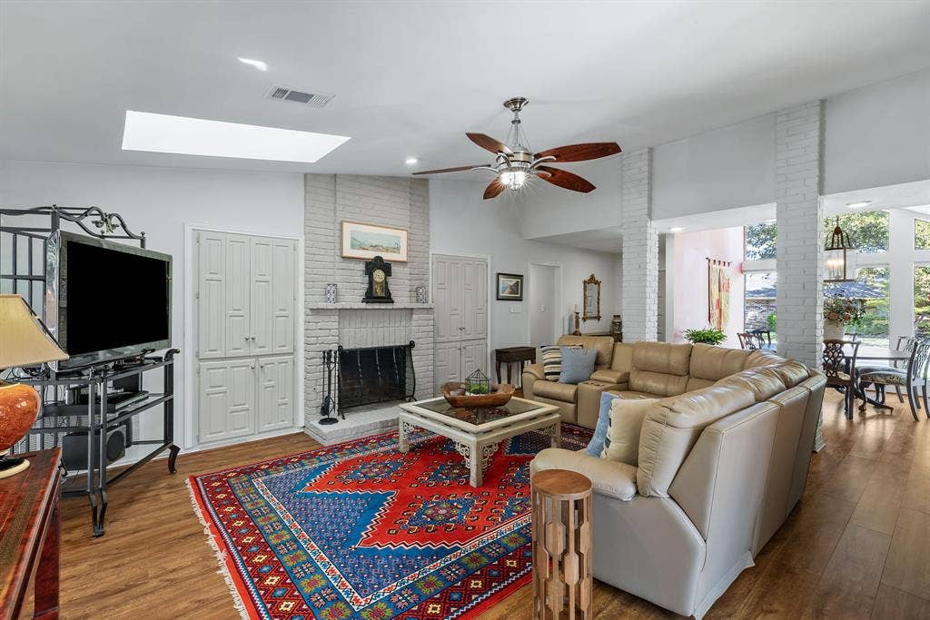 6803 Fernshaw Drive, Dallas, TX 75248 Vaulted ceiling with skylight, wood-finish flooring, white-painted brick fireplace, built-in cabinetry, and ceiling fan with lighting