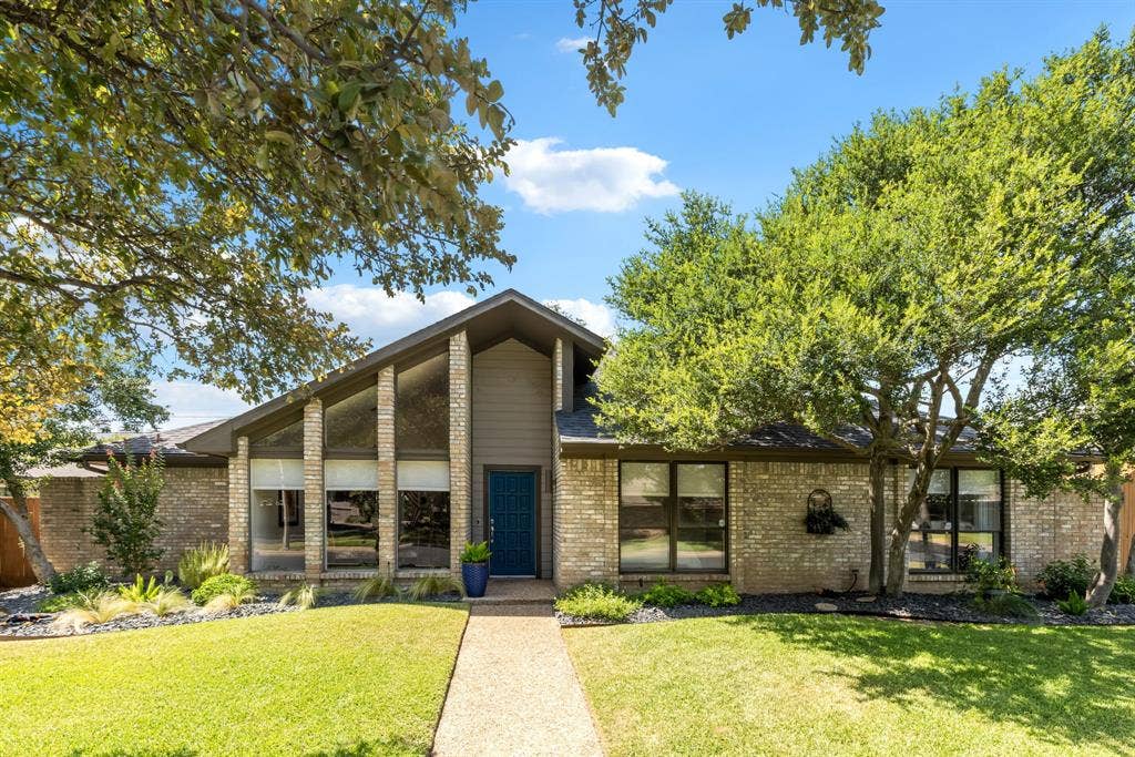 6803 Fernshaw Drive, Dallas, TX 75248 Brick exterior with a peaked roofline, a striking blue entry door, expansive front windows, mature trees, and a concrete walkway
