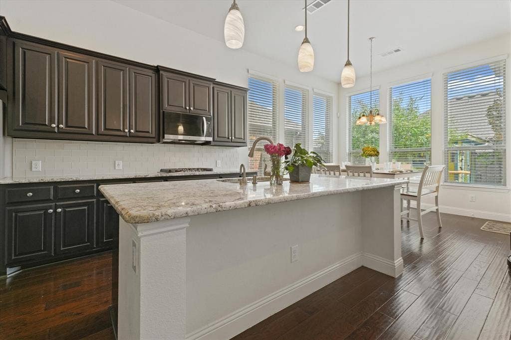 1113 5th Street, Argyle, TX 76226 Open kitchen with a large granite island, dark cabinetry, pendant lighting and an adjoining breakfast area surrounded by windows.