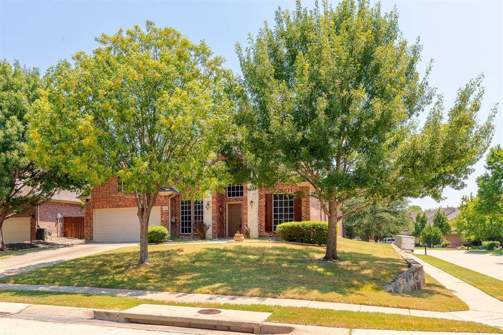 1913 Abilene Way, McKinney, TX 75072 Brick facade home featuring a two-car garage, mature front yard trees, a sloped lawn, and a stone retaining wall